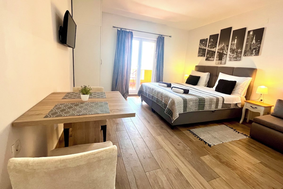 A modern hotel room with a wooden floor, a bed with a grey headboard, a bedside table with a lamp, a sofa, and a TV mounted on the wall.