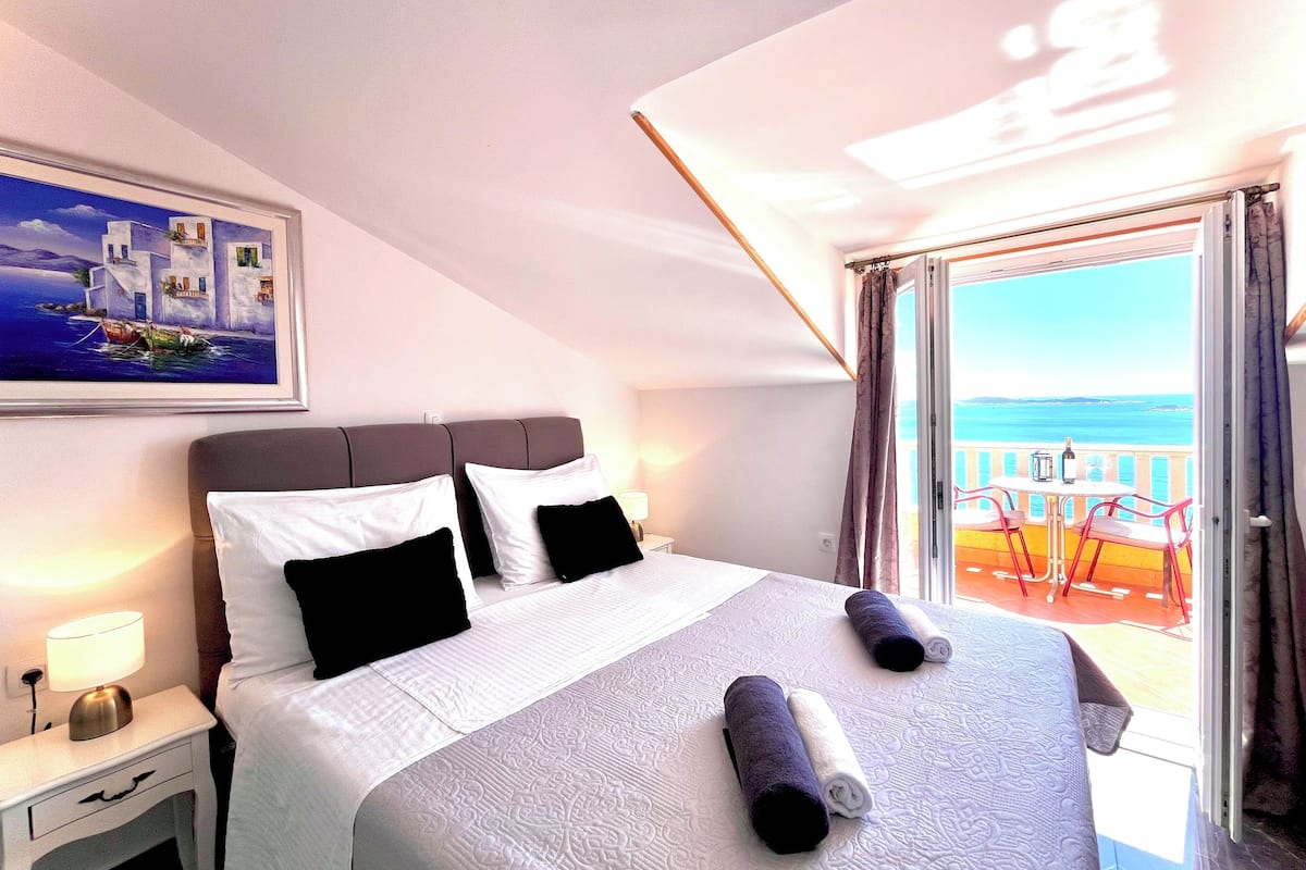 A hotel room with a bed, a nightstand, a painting of a coastal scene, and a balcony with a view of the sea.