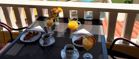 Ein Tisch reichhaltig gedeckt mit Croissants, Kaffee, Saft und einem Glas Orangensaft, mit Blick auf eine malerische Landschaft aus Meer und Grün.
