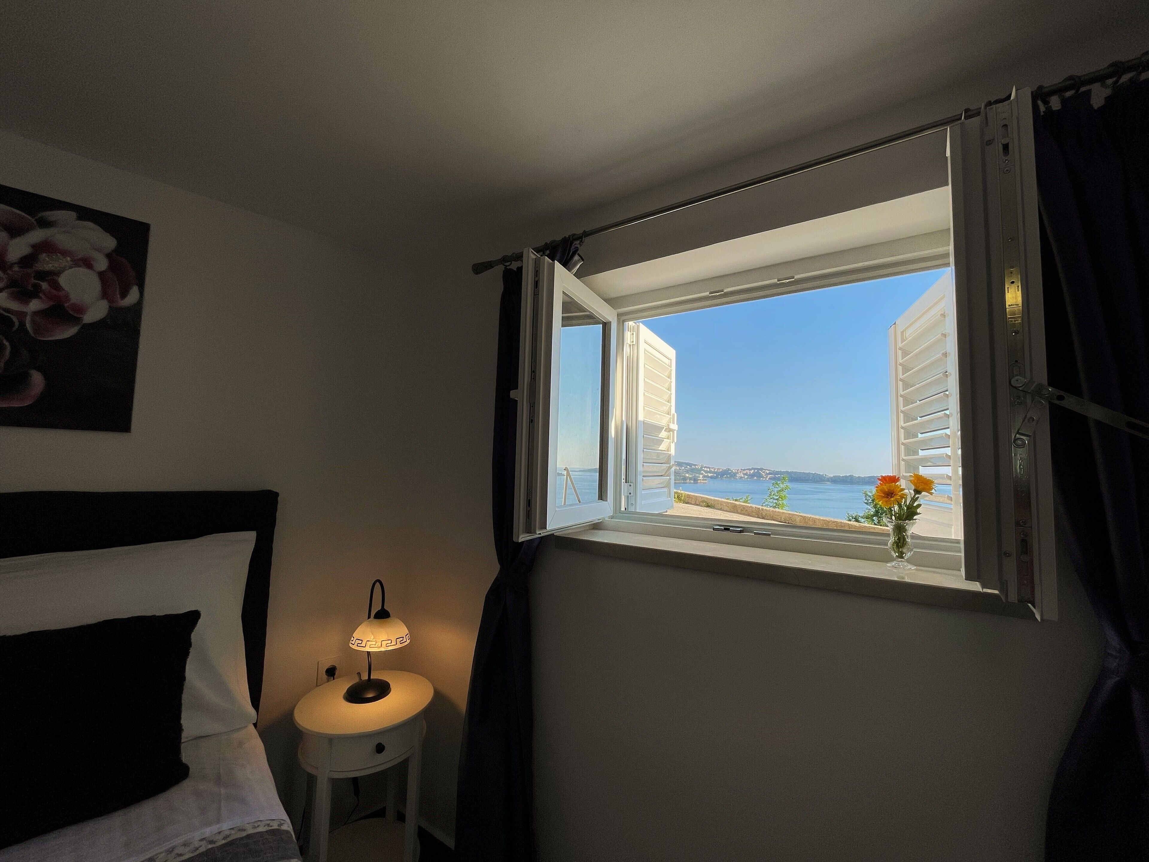 A hotel room with a bed, a bedside lamp, a window with curtains, and a view of the sea and buildings.