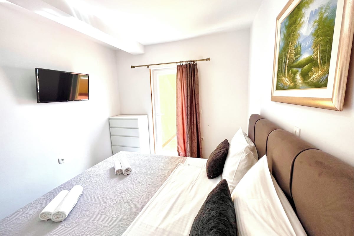 A hotel room with a bed, a flat-screen TV, a painting, and a window with curtains.