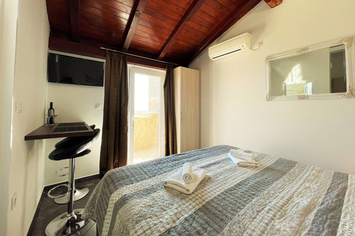 A bedroom with a bed, a desk with a chair, a TV mounted on the wall, and an air conditioning unit.