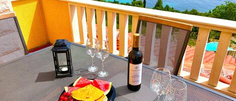 A table with a bottle of wine, glasses, and a plate with fruit on a balcony overlooking a scenic view of a lake and hills.