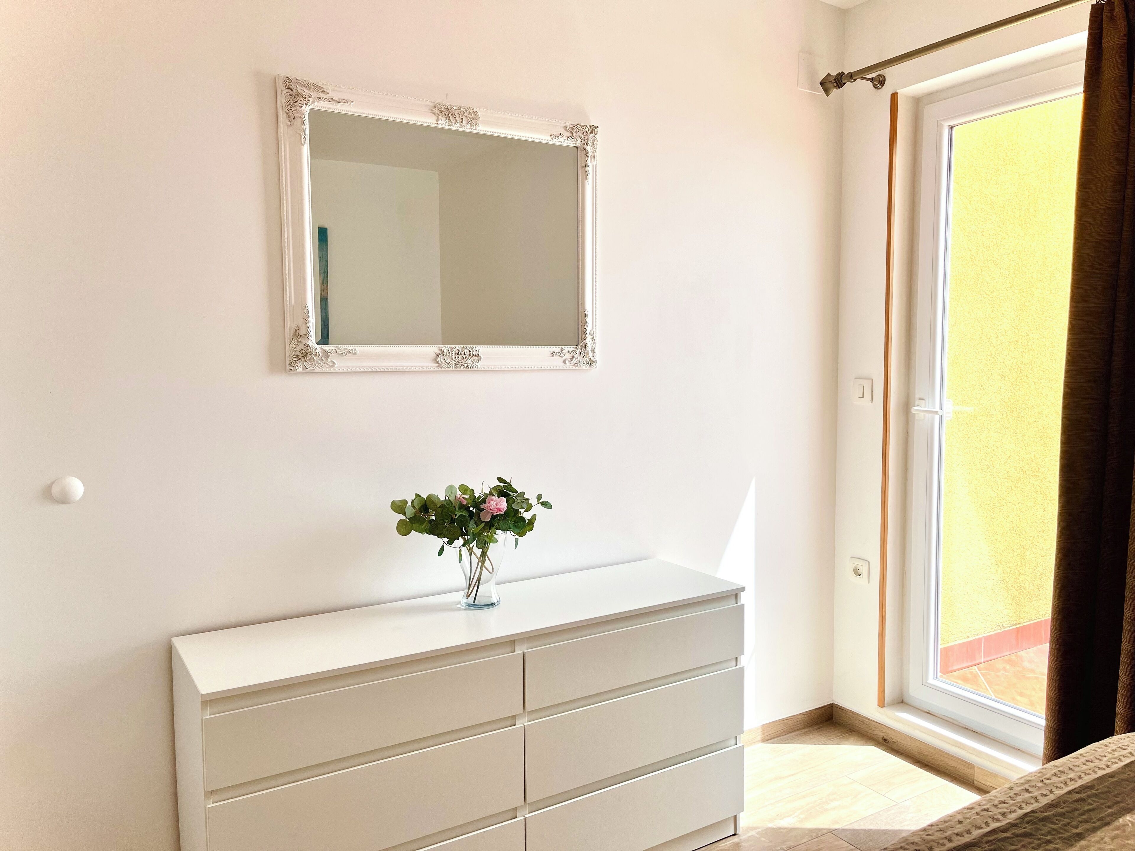 A bedroom with a white dresser, a mirror with an ornate frame, and a window with a view of the outside.
