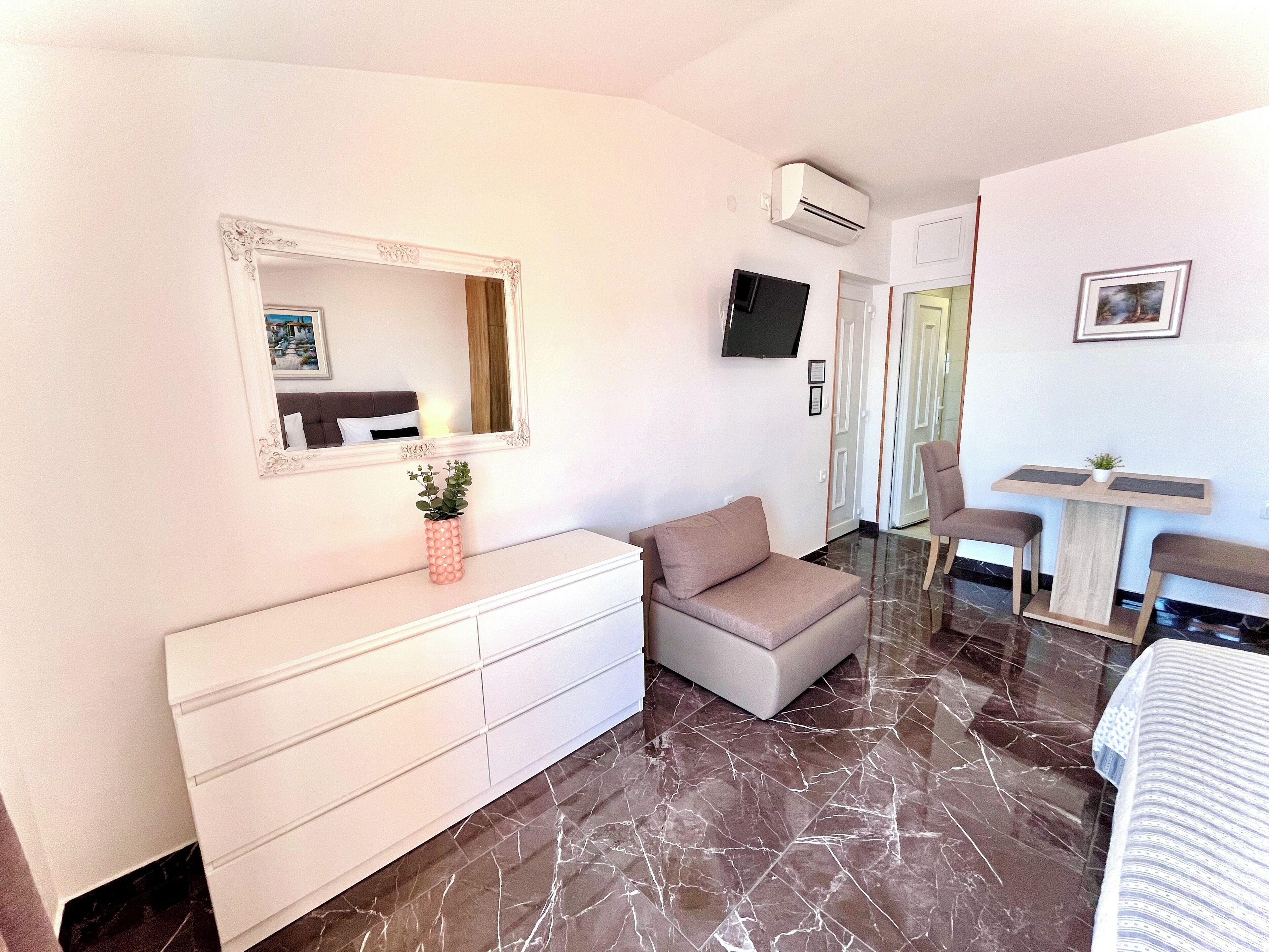 Un dormitorio moderno con un espejo grande, televisor, cómoda, sofá y una mesita con una planta.