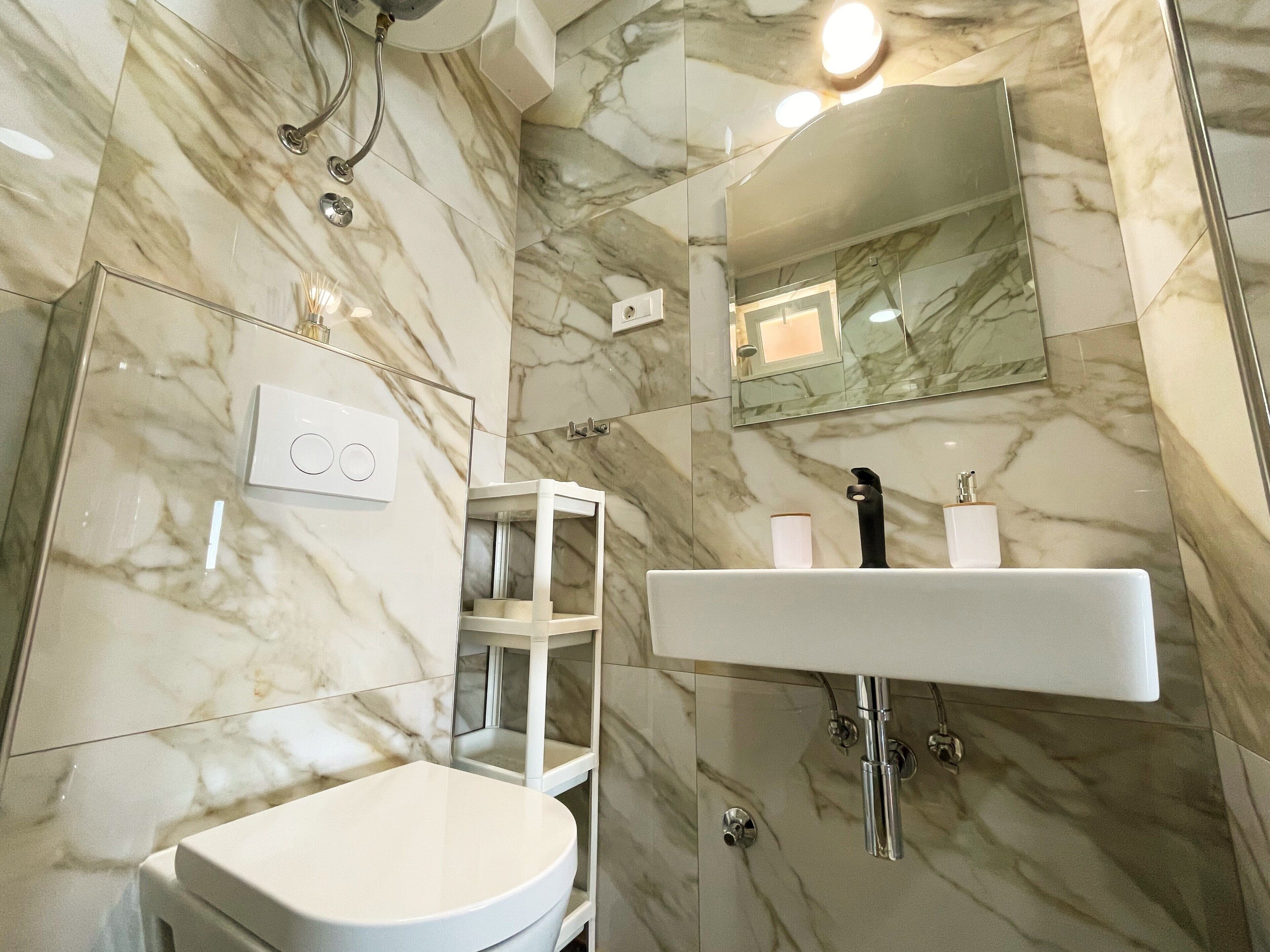 Une salle de bain moderne avec un sol en marbre, des toilettes blanches, une douche en verre et un lavabo blanc avec un miroir au-dessus.