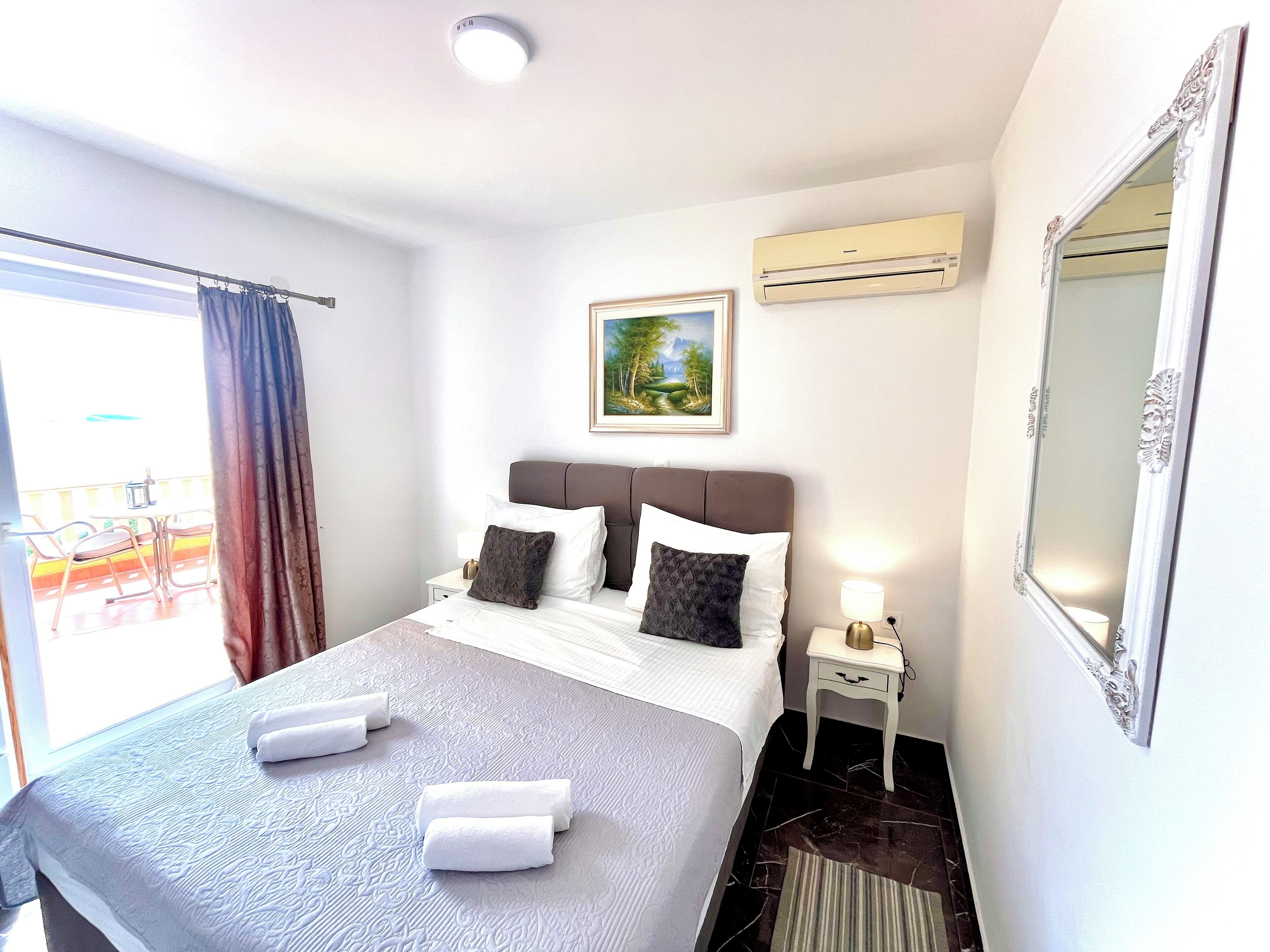 A hotel room with a bed, a bedside table, a nightstand, a mirror, an air conditioning unit, and a window with curtains.