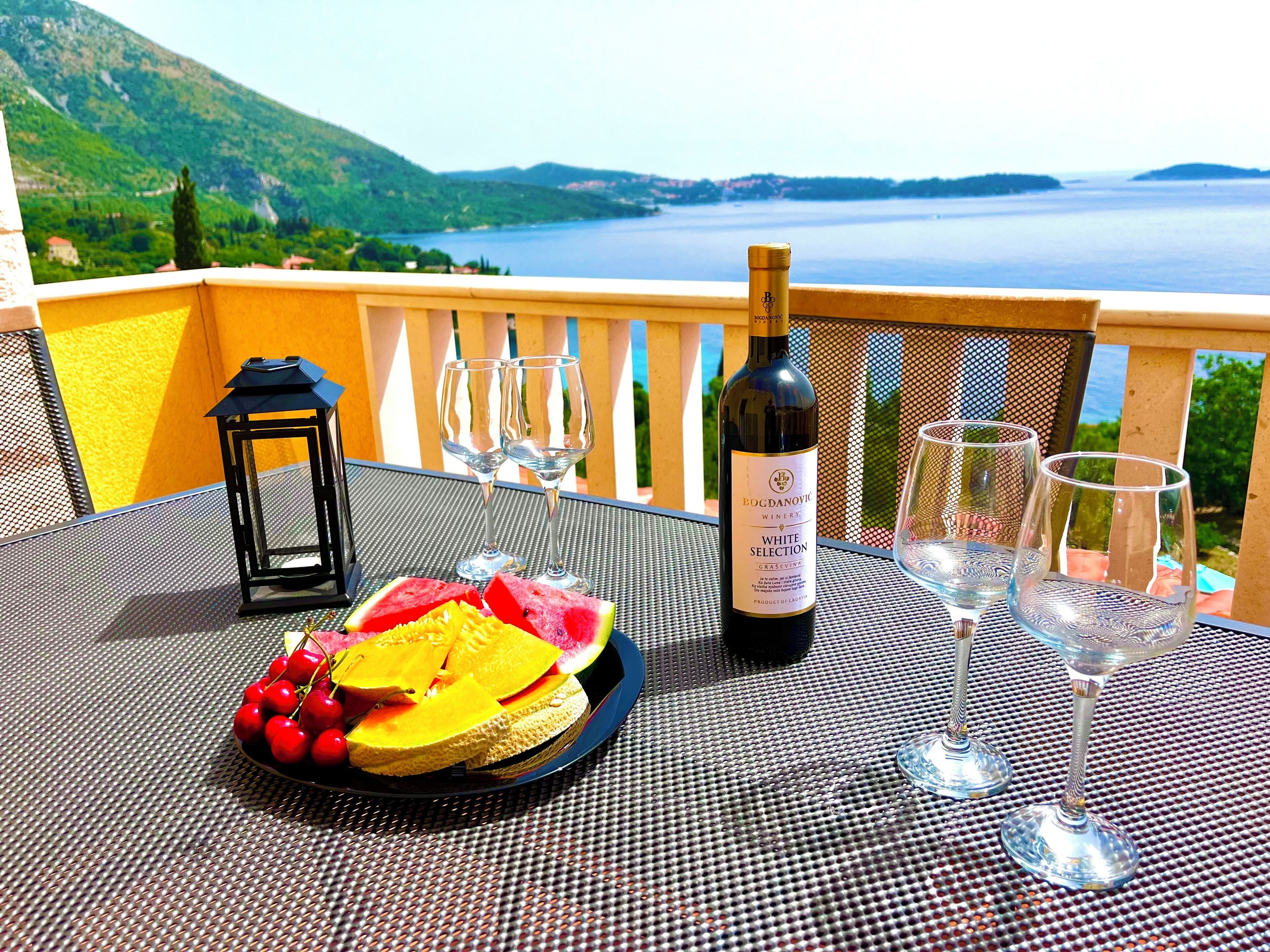 Una mesa con una botella de vino, copas, un farol y fruta en un balcón con vista a un lago.