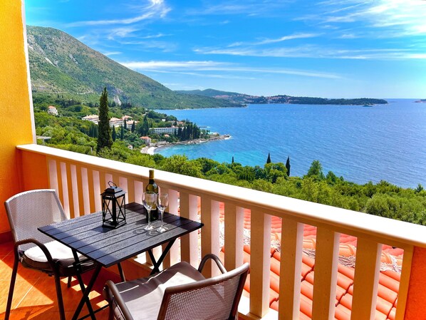A balcony with a table and chairs, overlooking a coastal landscape with hills and a body of water. - Villa Sunset (Zupa dubrovacka)