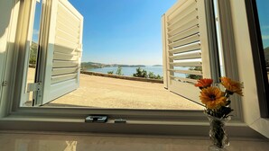 Una ventana con contraventanas abiertas, un jarrón con flores y vistas al paisaje costero.