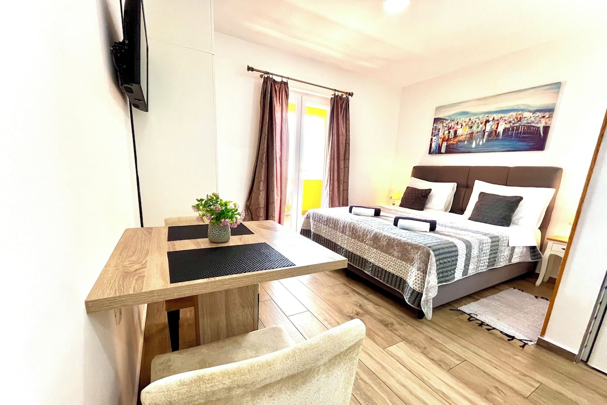 A hotel room with a bed, a desk with a chair, a small table with a plant, and a painting on the wall.