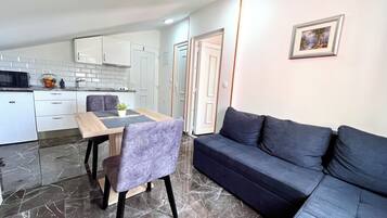 Una sala moderna con un sofá azul oscuro, una mesa de comedor de madera con dos sillas, y una zona de cocina con armarios blancos y piso de mármol.