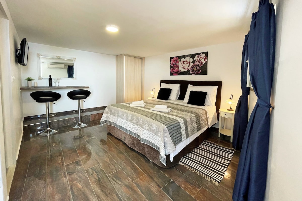 A hotel room with a bed, two bar stools, a small table with a mirror, and a wall-mounted TV.