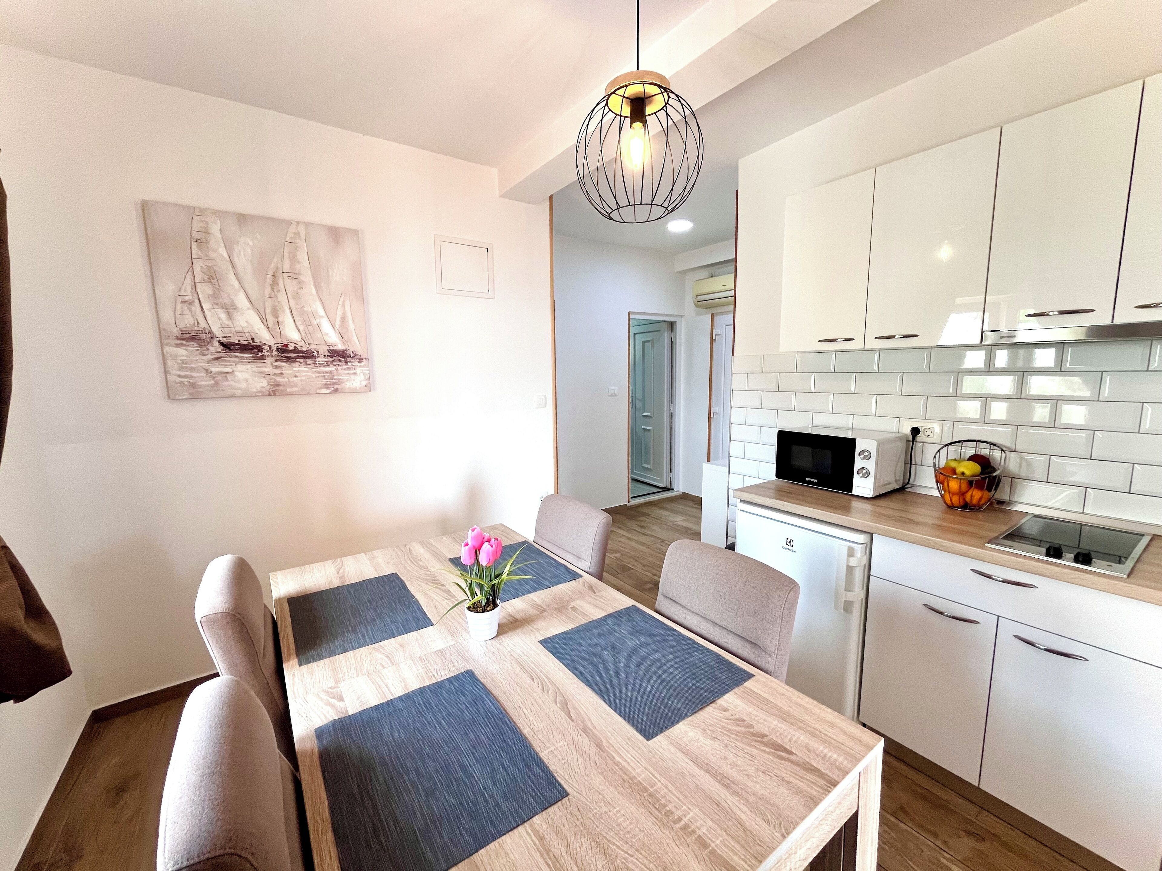 A modern kitchen with white cabinets, a microwave, and a sink. There is a dining area with a table and chairs, and a painting on the wall.