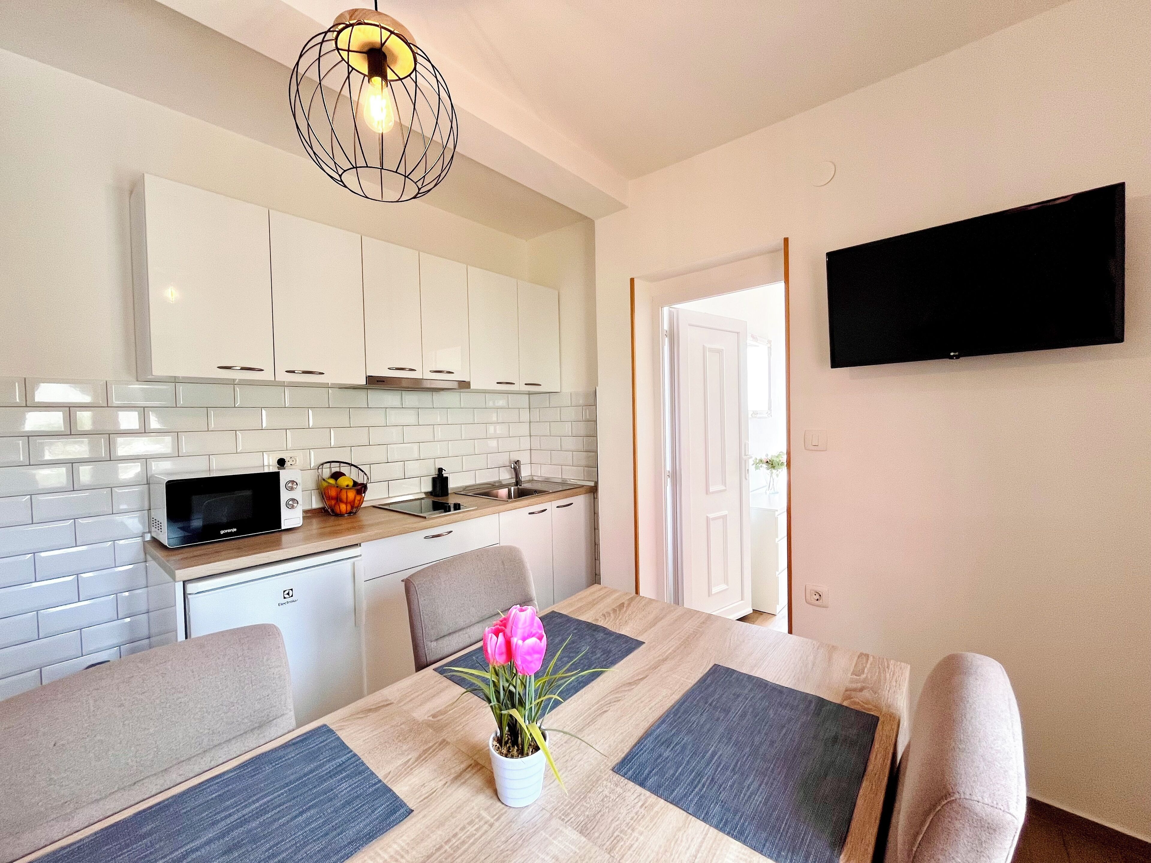 Cocina moderna con armarios blancos, un microondas y un frutero. Hay una mesa de comedor con individuales y un jarrón de flores. Un televisor de pantalla plana está montado en la pared.