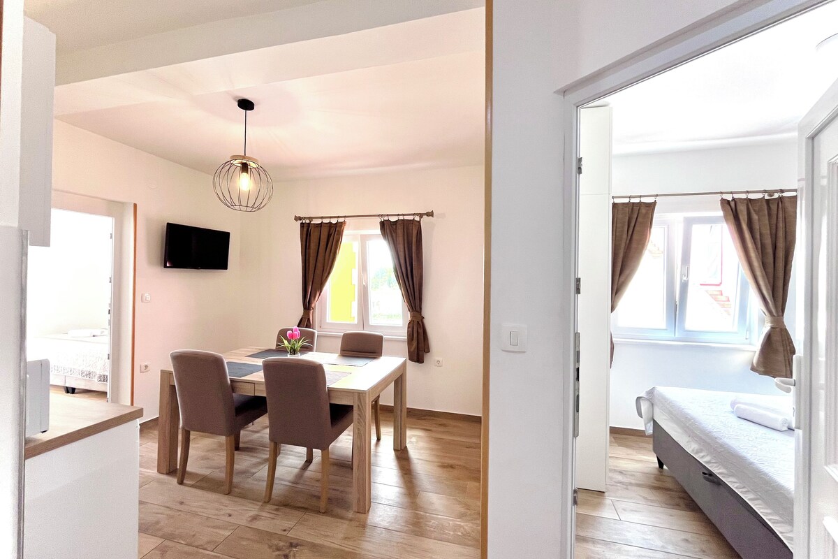 Una habitación moderna con suelo de madera, una mesa de comedor con sillas, un televisor de pantalla plana montado en la pared y una cama con cortinas.