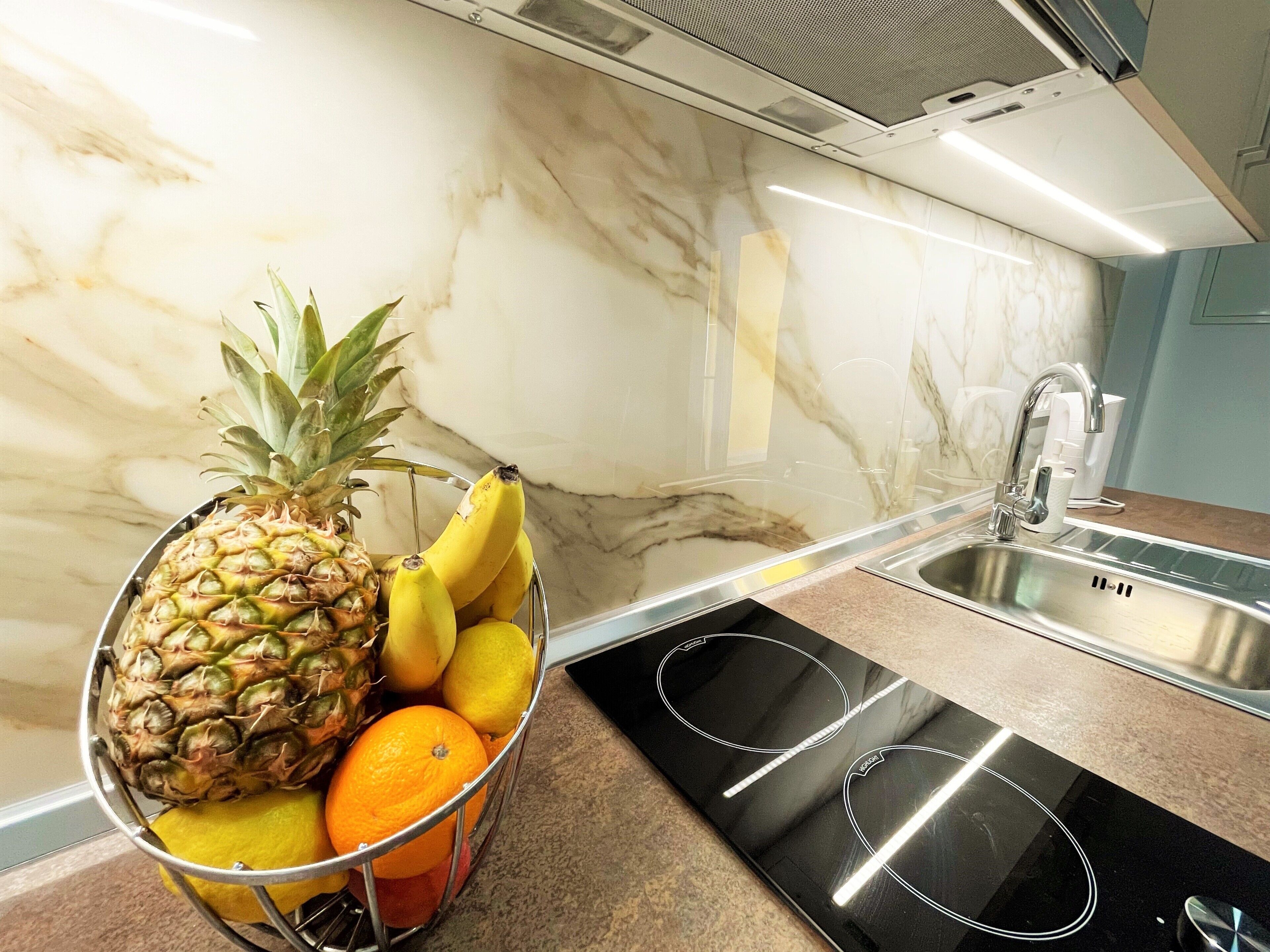 Cocina con salpicadero de mármol, lavamanos de acero inoxidable y frutero con piña, plátanos y naranjas.
