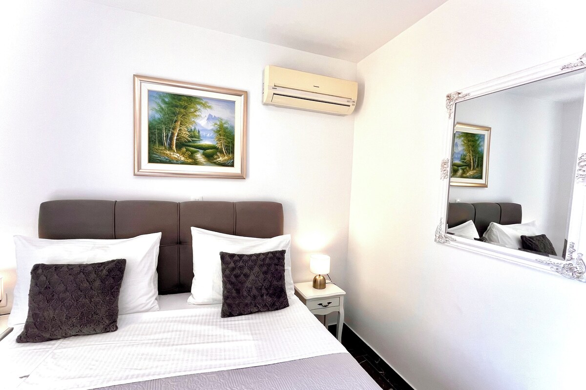 Habitación de hotel con cama, ropa de cama blanca, mesita de noche con lámpara, aire acondicionado y cuadros en la pared.