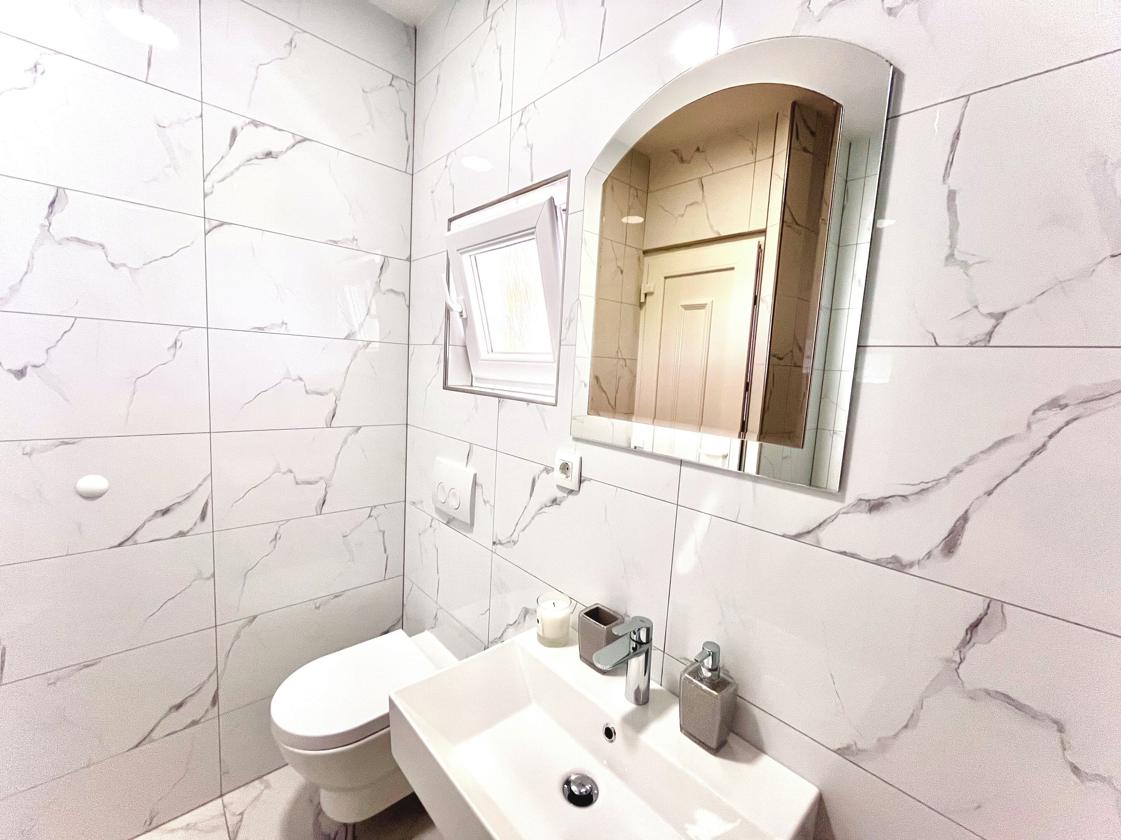 A bathroom with white marble tiles, a toilet, a sink with a mirror, and a shower area.