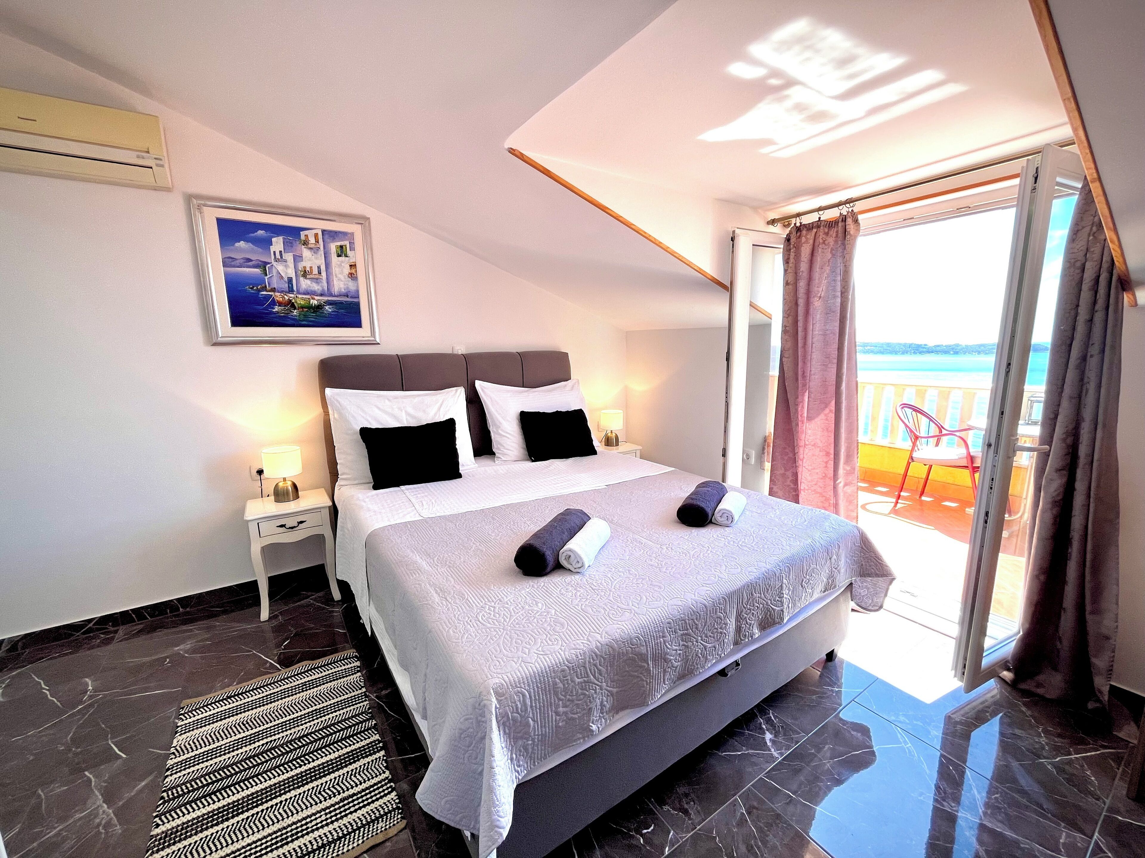 A hotel room with a bed, a nightstand, a lamp, a painting on the wall, and a view of the sea through a balcony.