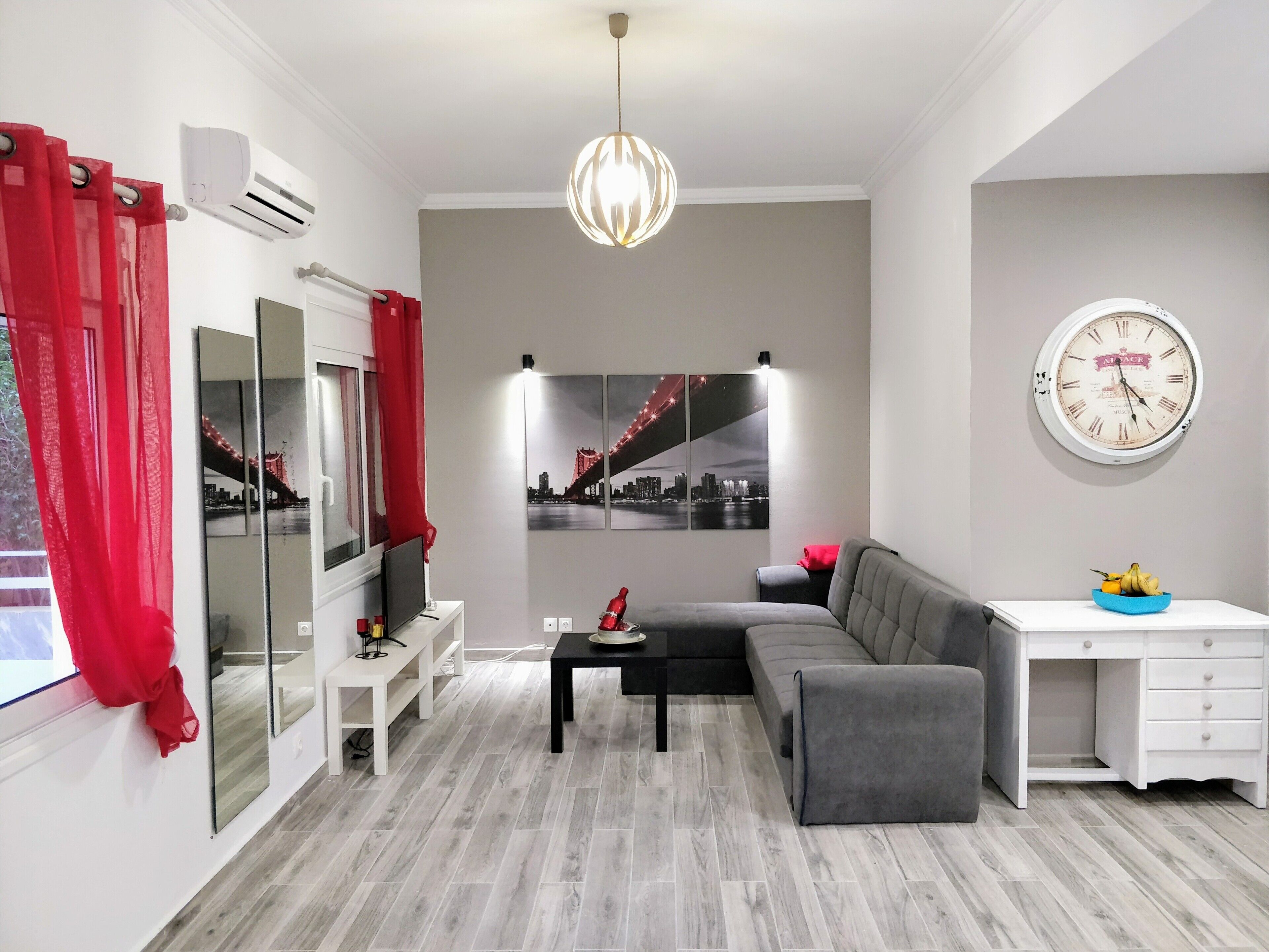 Ein modernes Wohnzimmer mit grauem Sofa, einem schwarzen Couchtisch, einem weißen Schreibtisch und einem großen Spiegel. Am Fenster hängt ein roter Vorhang und eine Stadtansicht ist als Gemälde an der Wand zu sehen.