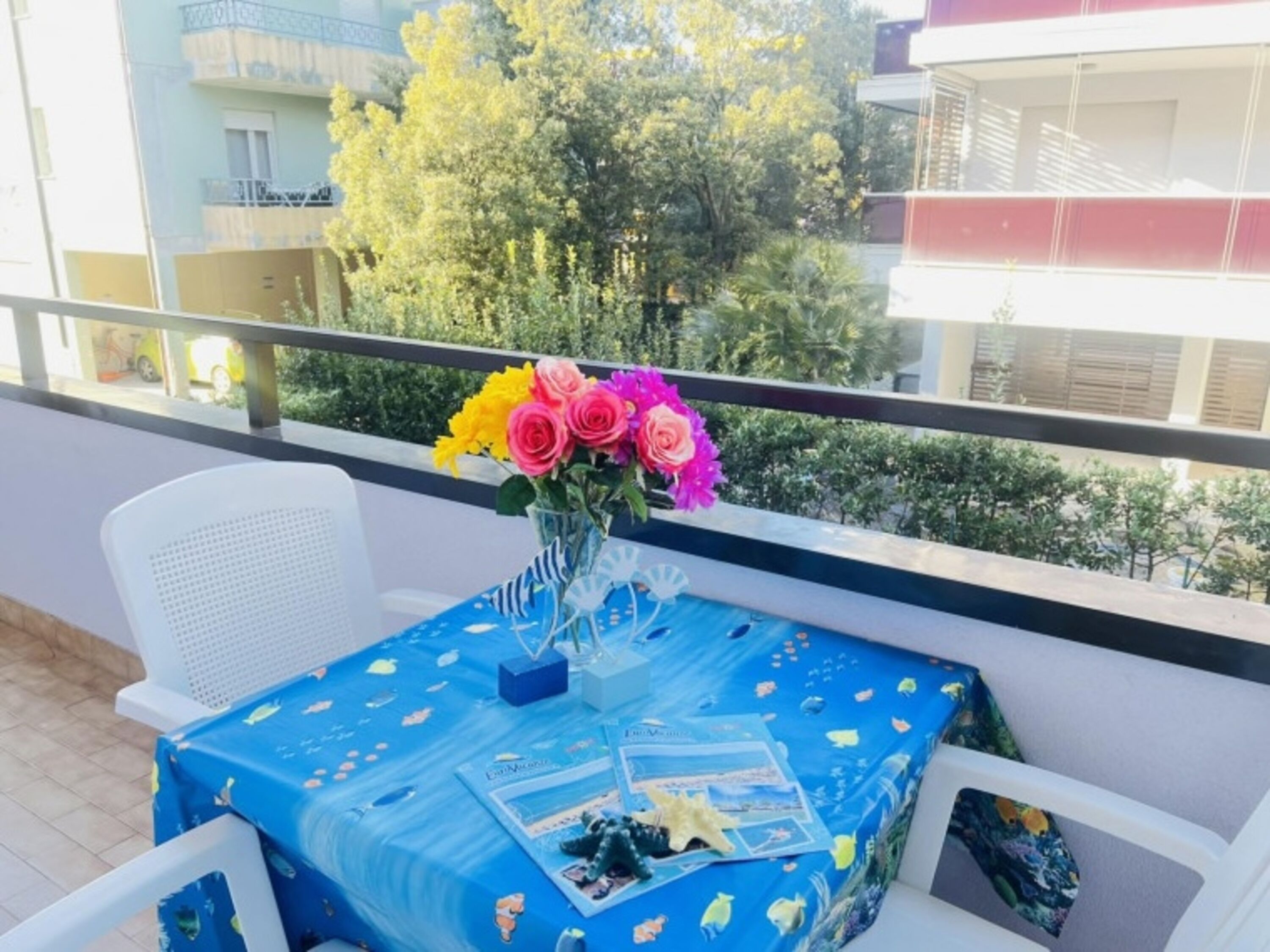 Un balcon avec une table recouverte d’une nappe bleue, une chaise blanche et un vase rempli de fleurs colorées.