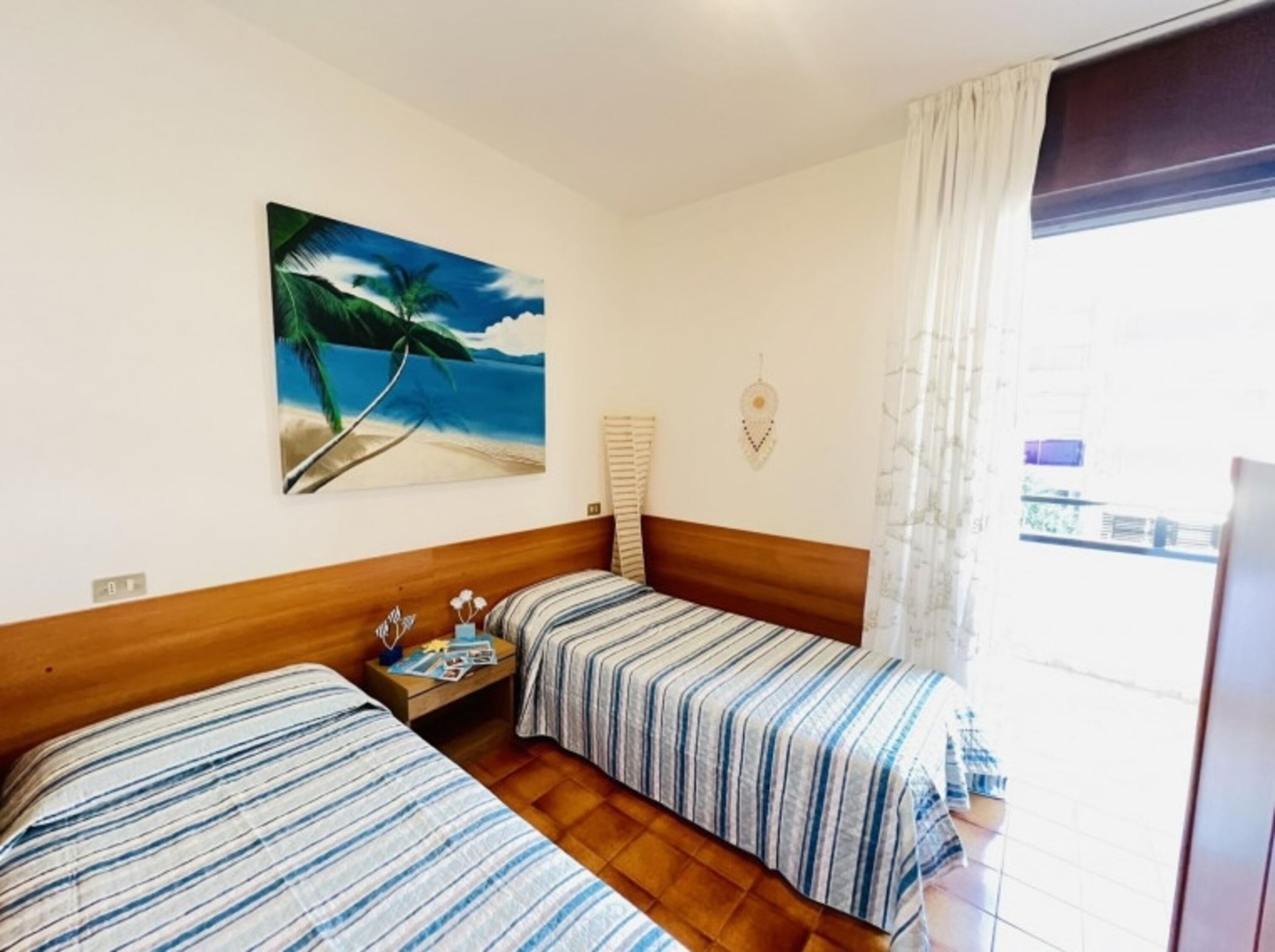 Ein Hotelzimmer mit zwei Betten, einem Strandbild, einem Fenster mit Vorhängen und einem hölzernen Kopfteil.