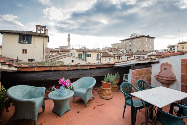 A terrace with wicker furniture, potted plants, and a view of historic buildings.