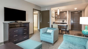Una habitación de hotel moderna con televisión de pantalla plana, un sofá, una silla, un área de comedor con mesa y sillas, y una cocina.