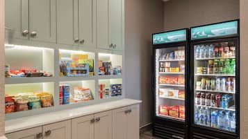 Una despensa de hotel con estantes llenos de bocadillos y bebidas, y dos refrigeradores con la etiqueta 'Homewood Suites'.
