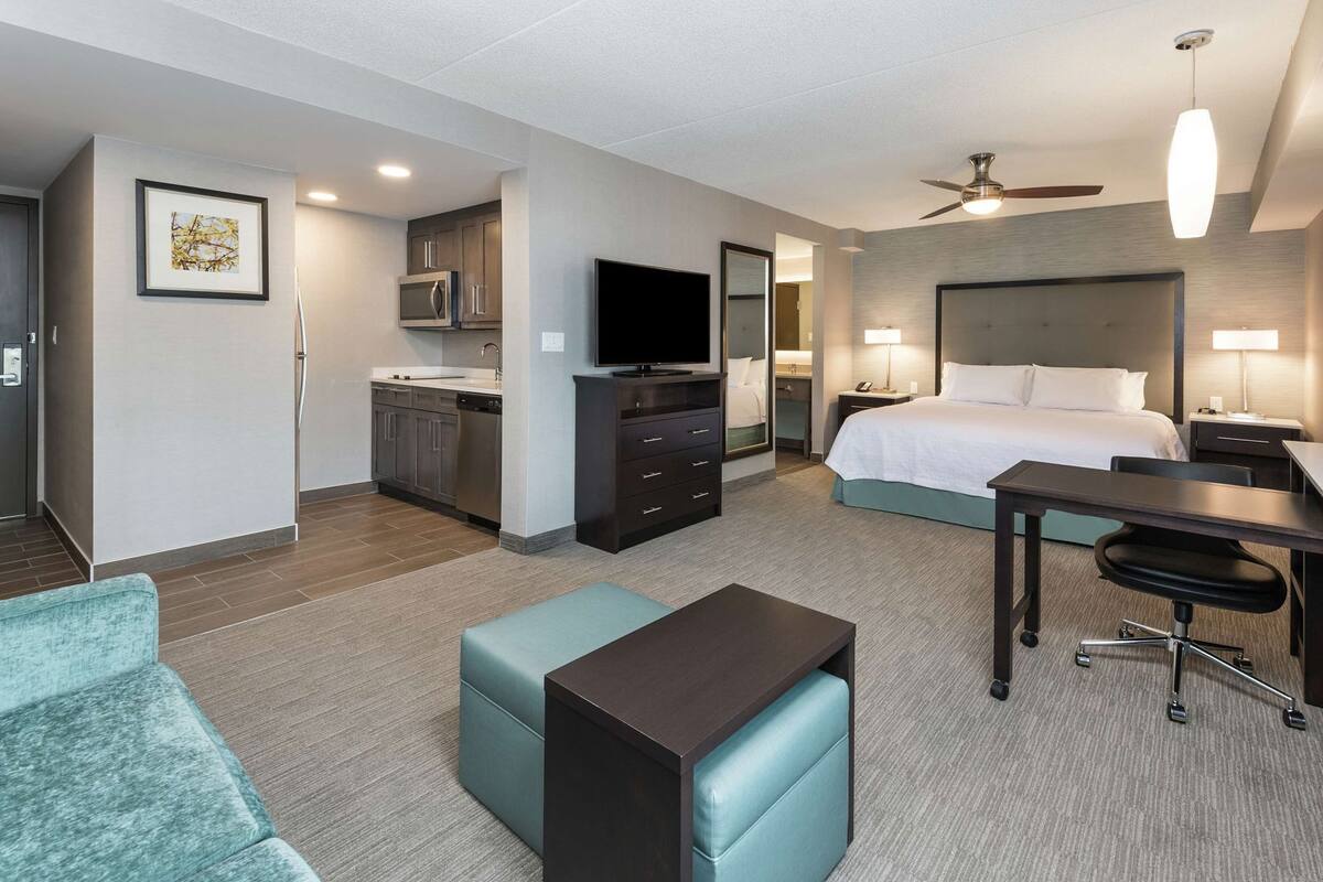 A hotel room with a kitchenette, a bed, a desk, a chair, and a ceiling fan.