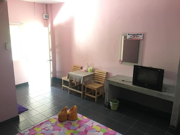 A small room with a tiled floor, a wooden table with chairs, a television on a stand, and a trash bin. - Mee Rak Resort (Bang Lamung)