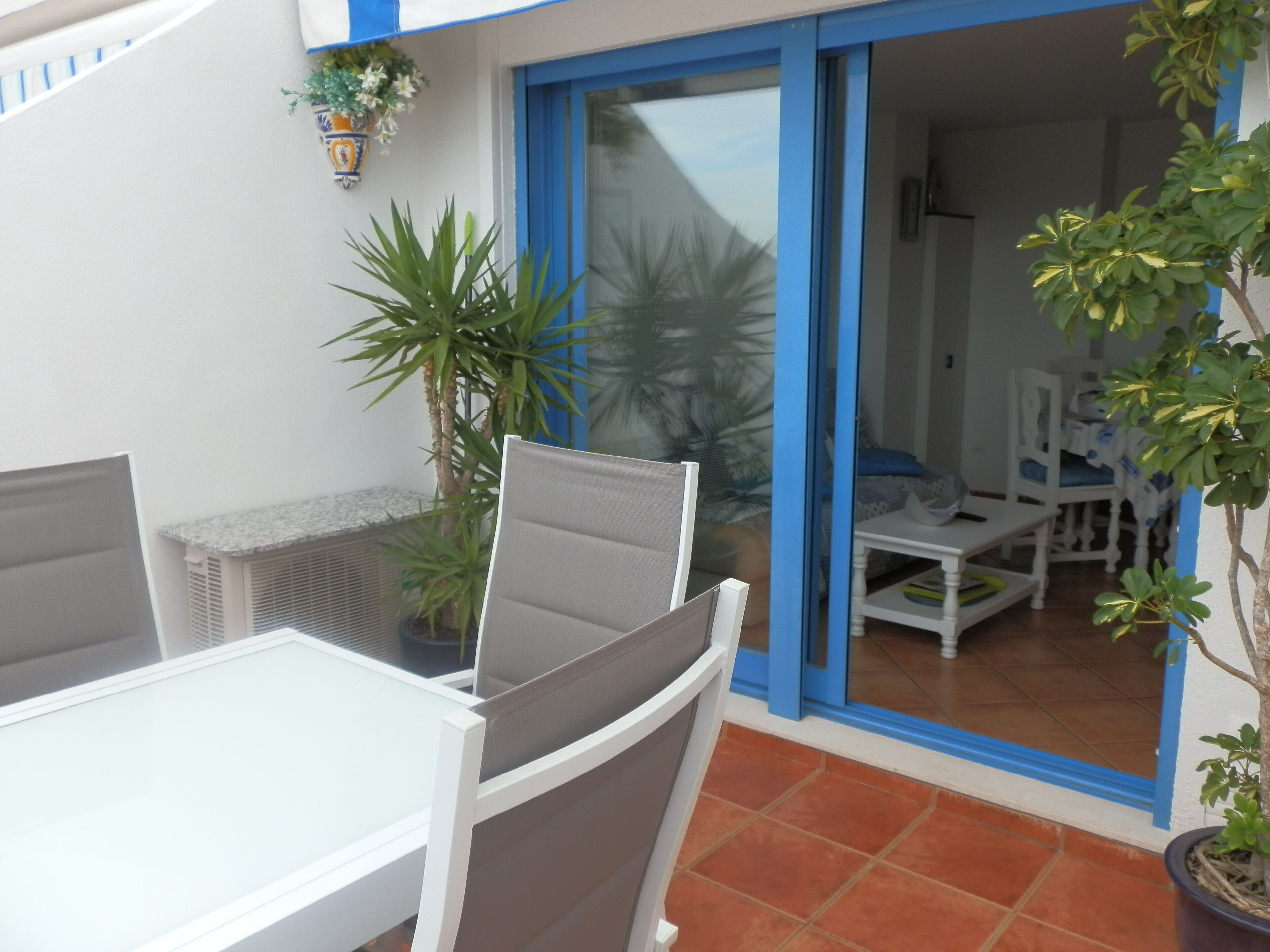 Eine Terrasse mit einem weißen Tisch und Stühlen, einem Topfpflanzen und einer blauen Tür, die zu einem Innenraum mit einem weißen Sessel und einem kleinen Tisch führt.