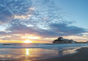 Un coucher de soleil sur la côte, avec un bâtiment historique perché sur une falaise dominant la mer.