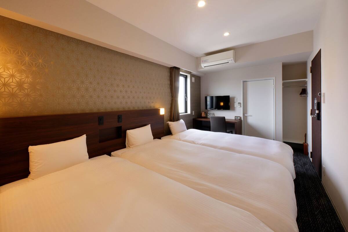 Quarto de hotel com duas camas grandes, uma escrivaninha e uma televisão, e um aparelho de ar condicionado.