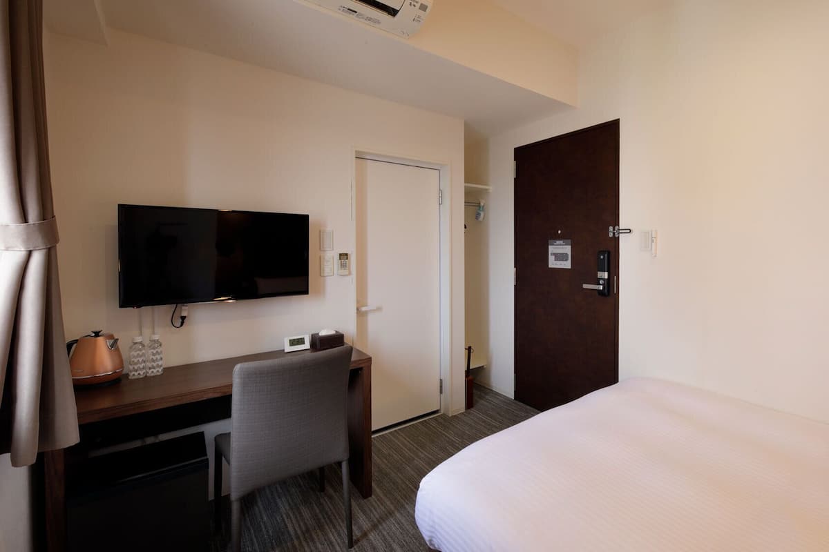 Ein Hotelzimmer mit Bett, Schreibtisch mit Stuhl, Fernseher, Tür und einer Wand-Klimaanlage.