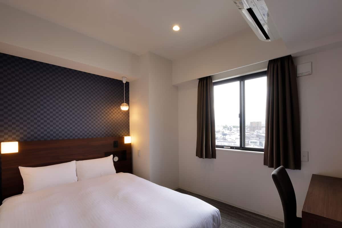 Ein Hotelzimmer mit einem großen Bett, einem Schreibtisch, einem Stuhl, einem Fenster mit Vorhängen und Blick auf die Stadt.