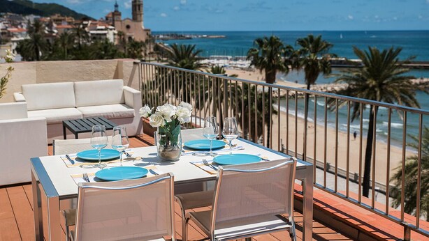 Une terrasse avec une table dressée pour deux personnes, offrant une vue sur une plage et un bâtiment historique.