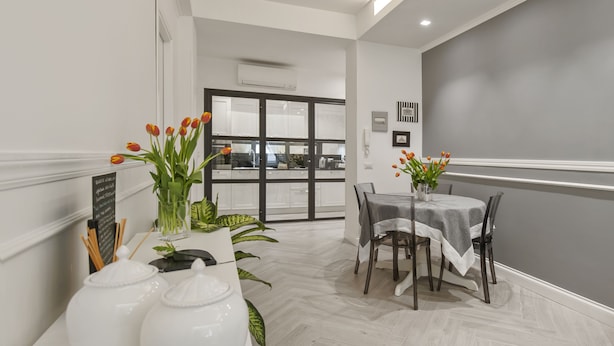 Un espace repas moderne avec une table blanche, des chaises et des tulipes. On aperçoit une fenêtre avec des stores et une porte au fond.