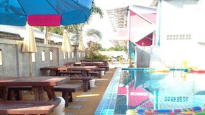 Uma área junto à piscina com mesas e bancos de madeira, piscina azulejada e guarda-sóis.