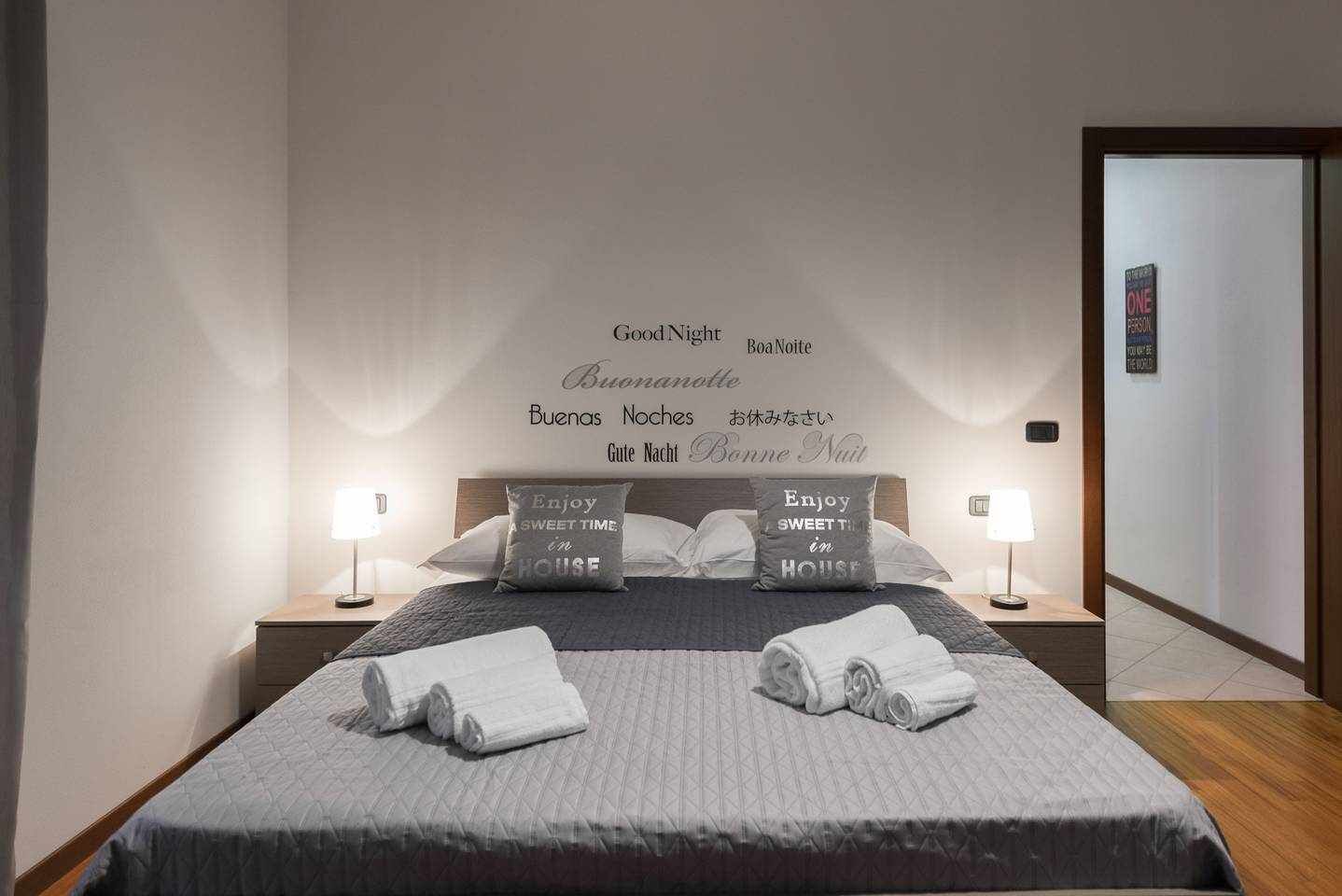 Un lit bien fait, avec des oreillers, une table de chevet avec une lampe et un mur orné de messages multilingues souhaitant une « bonne nuit ».