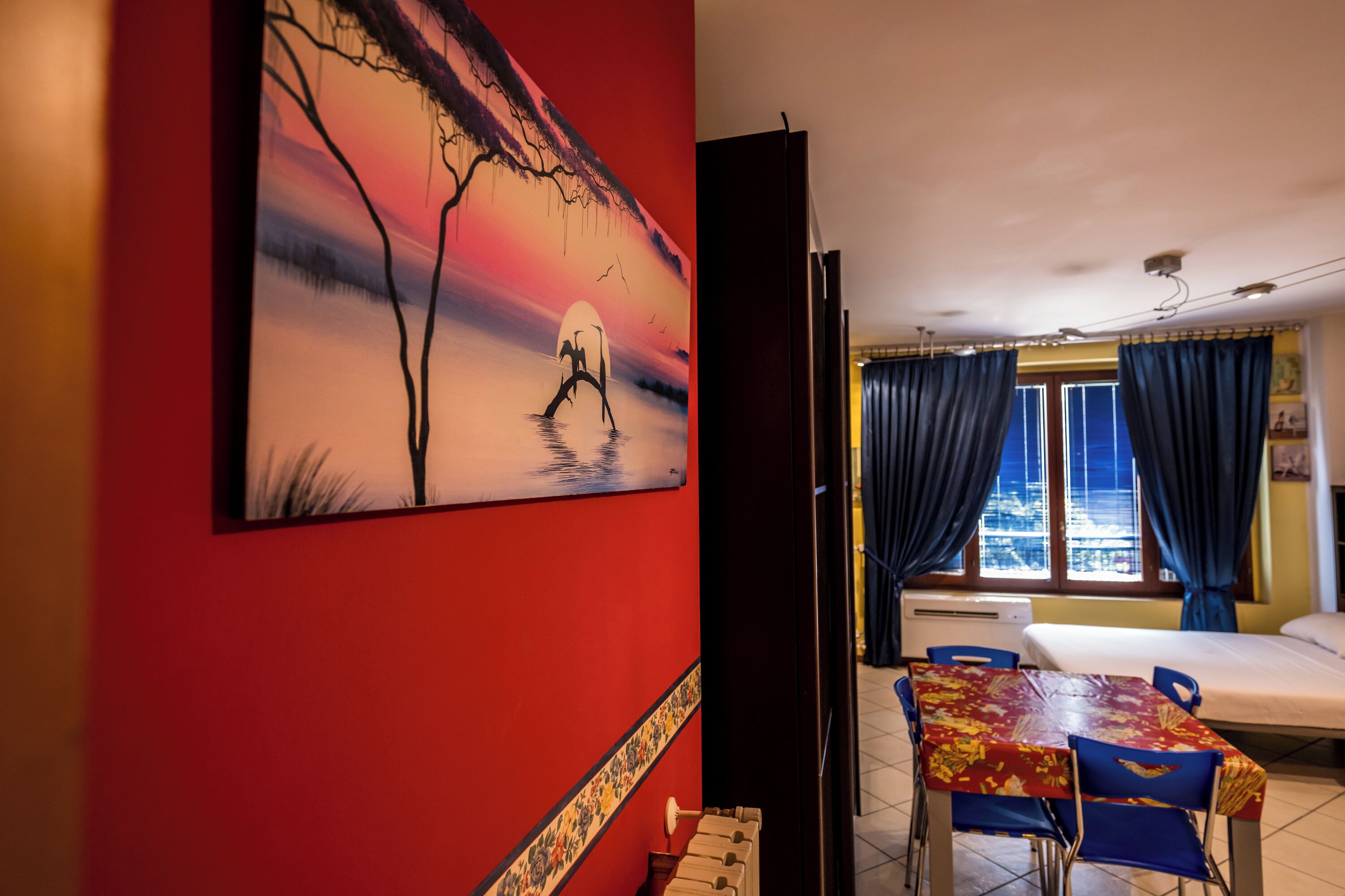 A hotel room with a red wall, a painting of a sunset, a bed, a table with chairs, and a window with curtains.