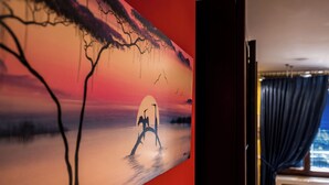 A room with a large painting of a sunset scene on the wall, a window with curtains, and a visible air conditioning unit. - Cozy Suite Arena Milano Santa Giulia (MILANO)