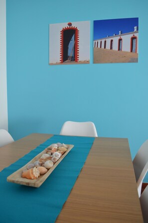 Une table à manger recouverte d’une nappe bleue, avec des chaises blanches et un plateau en bois orné de coquillages.