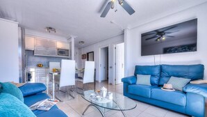 Una sala de estar moderna con un sofá azul, una mesa de centro de vidrio y una cocina al fondo.