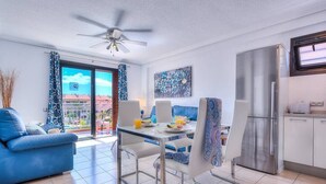 Una sala moderna con un sofá azul, una mesa de comedor con sillas, un ventilador de techo, un ventanal con cortinas y una zona de cocina con refrigerador.