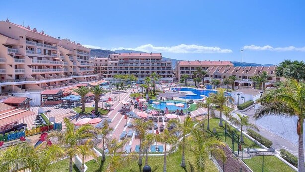 Un resort con varias construcciones, una zona de piscina central y palmeras.