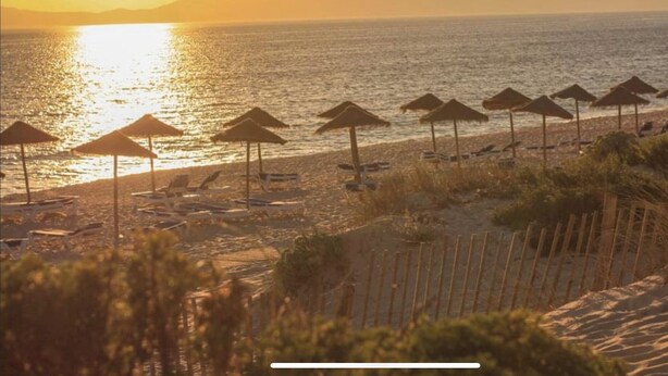 A beach with sun loungers and thatched umbrellas, a sandy shoreline, and a sunset over the ocean.