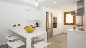 Une cuisine moderne avec des meubles blancs, un réfrigérateur en acier inoxydable, un four et un four à micro-ondes. On y trouve une table à manger avec des chaises blanches et un saladier rempli de fruits.