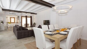 Un espace repas moderne avec une table en bois dressée pour six personnes, entourée de chaises blanches. Un canapé foncé et une table à manger avec un plateau en verre sont visibles au fond.